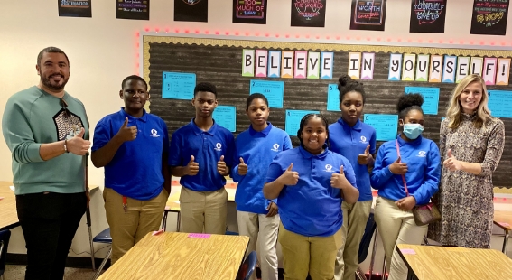 Six students and two teachers in a classroom giving a thumbs-up signal