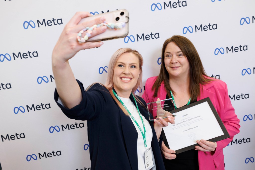 Two women taking a selfie in front of a Meta logo wall.